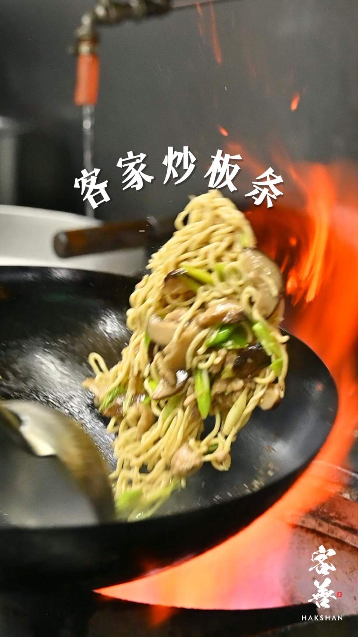 Yellow Hakka stir-fried egg noodles airborne above a carbon-steel wok over a bright orange flame