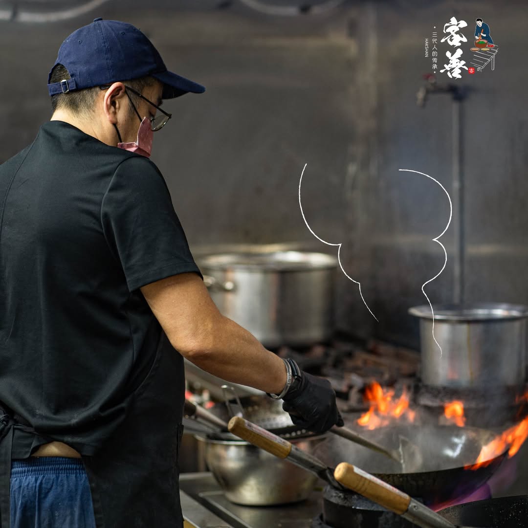A Hakshan chef in a cap and dark apron working at the open-kitchen wok station, flame visible