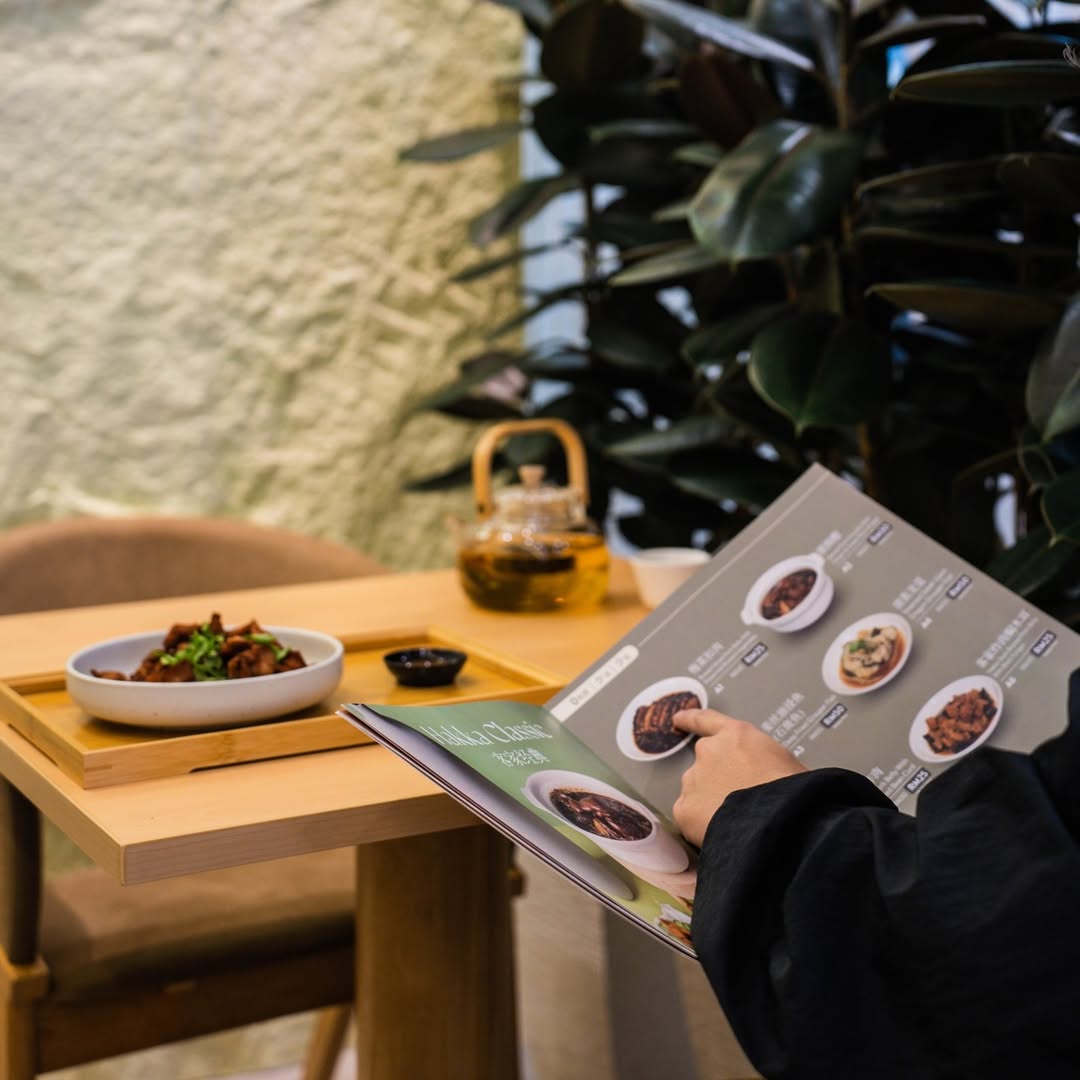 A guest at a Hakshan table reading the menu booklet over a small Hakka dish, chopsticks, and sauces