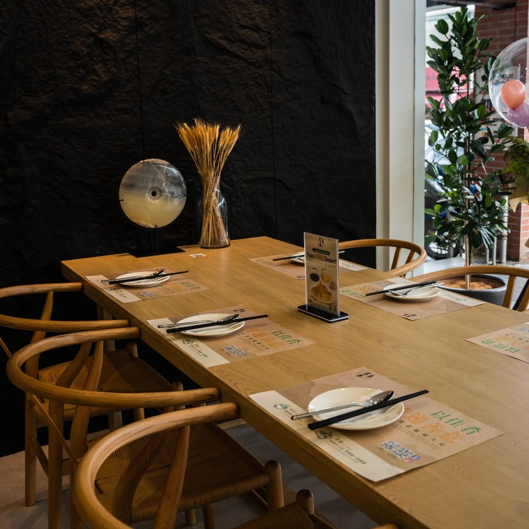 A long oak Hakshan dining table set for shared dining, surrounded by wishbone chairs, with a dried wheat spray on the wall ledge